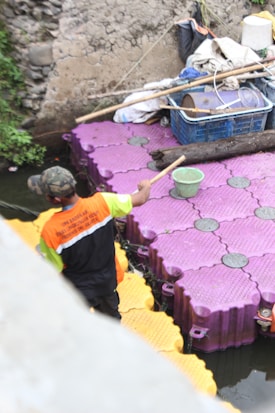 A person in a bright orange and yellow vest stands on floating modular platforms over a body of water. The platforms are purple and yellow. Nearby, there is a basket filled with various items, including sticks and a drum. The scene is set against a stone wall, and there is some greenery on one side.