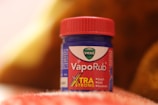A blue and red container of Vicks VapoRub sits on a soft, textured surface. The label indicates it is an extra strong formulation for relief from coughs, colds, and headaches. The background has warm, out-of-focus hues.