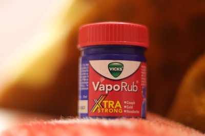 A blue and red container of Vicks VapoRub sits on a soft, textured surface. The label indicates it is an extra strong formulation for relief from coughs, colds, and headaches. The background has warm, out-of-focus hues.