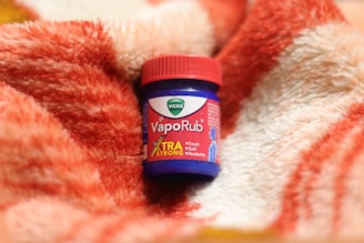 A jar of Vicks VapoRub extra strong variant is placed on a soft, plush blanket with a red and white pattern. The details on the jar indicate it's for cough, cold, and headache relief.