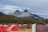 A series of red tents are set up in a grassy camping area with wooden dividers. In the background, there are majestic, rocky mountains under a cloudy sky. Two people are sitting at a picnic table near the tents.