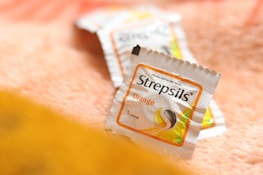 A close-up of Strepsils lozenge packaging, positioned on a soft, textured surface with a warm color tone. The packaging is white with an orange border, indicating it's an orange-flavored variant.