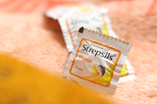 A close-up of Strepsils lozenge packaging, positioned on a soft, textured surface with a warm color tone. The packaging is white with an orange border, indicating it's an orange-flavored variant.