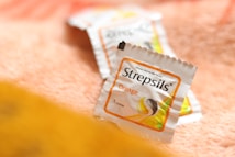 A close-up of Strepsils lozenge packaging, positioned on a soft, textured surface with a warm color tone. The packaging is white with an orange border, indicating it's an orange-flavored variant.