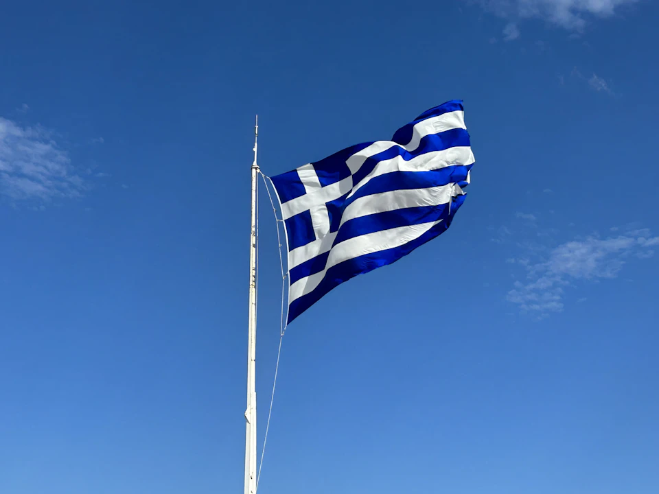 Greek flag flies high against a clear blue sky with white clouds