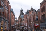 A picturesque street lined with historical red brick buildings featuring ornate architectural details. In the background, a prominent clock tower rises above the buildings. The street appears busy with people walking and a bus moving down the street. Several shop signs are visible, including one for Prezzo.
