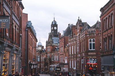 A picturesque street lined with historical red brick buildings featuring ornate architectural details. In the background, a prominent clock tower rises above the buildings. The street appears busy with people walking and a bus moving down the street. Several shop signs are visible, including one for Prezzo.