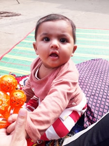 A baby dressed in a pink shirt is sitting on patterned mats outdoors, holding an orange toy. The baby is looking towards the camera with a curious expression. The background includes multicolored textiles, adding a vibrant touch.
