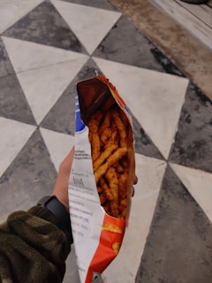 A hand is holding an open bag of snacks, revealing what appear to be spicy, crunchy sticks. The background consists of a geometric black and white tiled floor.