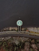 An aerial view shows a circular structure resembling a drain or spillway at the edge of a body of water. The structure is connected to a road by a narrow pathway that cuts through a rocky and vegetated area. The surrounding landscape includes patches of trees and bushes, with the water occupying the majority of the image.