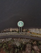 An aerial view shows a circular structure resembling a drain or spillway at the edge of a body of water. The structure is connected to a road by a narrow pathway that cuts through a rocky and vegetated area. The surrounding landscape includes patches of trees and bushes, with the water occupying the majority of the image.
