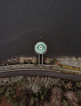 An aerial view shows a circular structure resembling a drain or spillway at the edge of a body of water. The structure is connected to a road by a narrow pathway that cuts through a rocky and vegetated area. The surrounding landscape includes patches of trees and bushes, with the water occupying the majority of the image.
