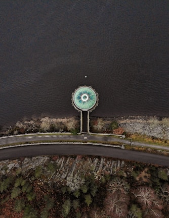 An aerial view shows a circular structure resembling a drain or spillway at the edge of a body of water. The structure is connected to a road by a narrow pathway that cuts through a rocky and vegetated area. The surrounding landscape includes patches of trees and bushes, with the water occupying the majority of the image.