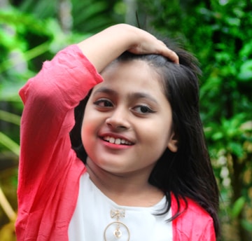 A young child with long hair is smiling while holding a hand over their head. They are wearing a white top with a decorative element and a bright pink jacket. The background is lush and green, suggesting an outdoor setting.