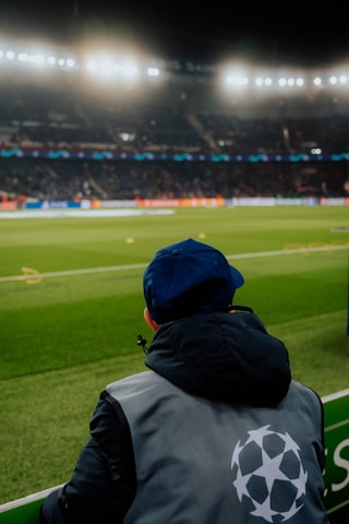 A person wearing a jacket with a logo stands near the edge of a brightly lit soccer field, gazing at the stadium filled with spectators under the floodlights.