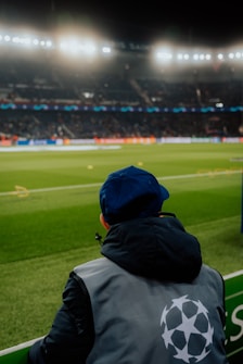 A person wearing a jacket with a logo stands near the edge of a brightly lit soccer field, gazing at the stadium filled with spectators under the floodlights.