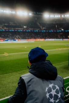 A person wearing a jacket with a logo stands near the edge of a brightly lit soccer field, gazing at the stadium filled with spectators under the floodlights.