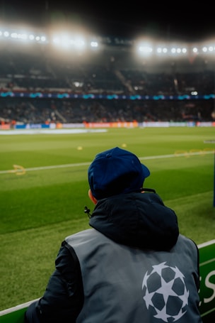 A person wearing a jacket with a logo stands near the edge of a brightly lit soccer field, gazing at the stadium filled with spectators under the floodlights.