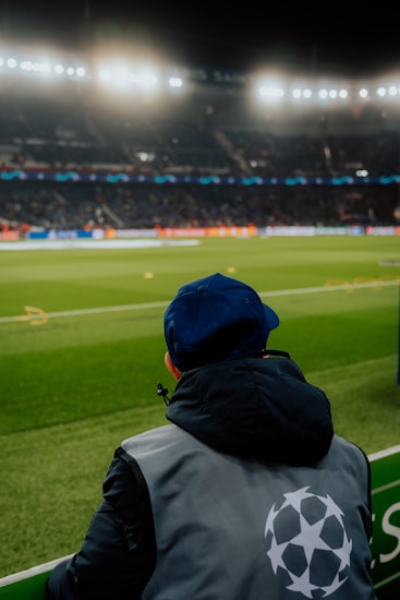 A person wearing a jacket with a logo stands near the edge of a brightly lit soccer field, gazing at the stadium filled with spectators under the floodlights.