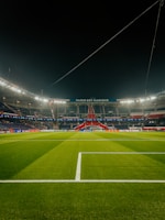 A well-lit football stadium with green grass, empty stands, and a prominent banner reading 'PARIS EST MAGIQUE' under the nighttime sky. The focus is on the field and the central structure in the background, which is decorated in red, white, and blue colors.