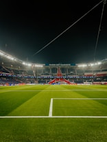 A well-lit football stadium with green grass, empty stands, and a prominent banner reading 'PARIS EST MAGIQUE' under the nighttime sky. The focus is on the field and the central structure in the background, which is decorated in red, white, and blue colors.