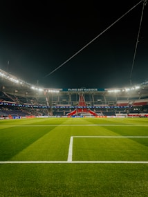 A well-lit football stadium with green grass, empty stands, and a prominent banner reading 'PARIS EST MAGIQUE' under the nighttime sky. The focus is on the field and the central structure in the background, which is decorated in red, white, and blue colors.