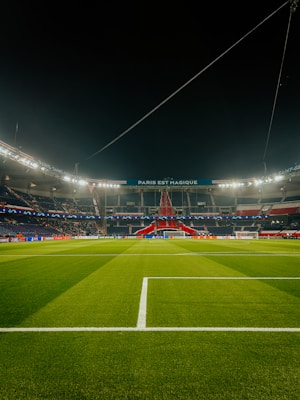 A well-lit football stadium with green grass, empty stands, and a prominent banner reading 'PARIS EST MAGIQUE' under the nighttime sky. The focus is on the field and the central structure in the background, which is decorated in red, white, and blue colors.