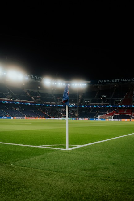 A well-lit empty football stadium at night, with a focus on one of the corner flags on the green field. Stadium lights illuminate the seating areas and advertisements are visible along the edges of the field. Banners with text can be seen around the stands.