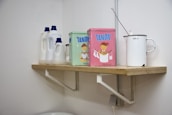 A wooden shelf is mounted on a white wall, holding several items. There are three white plastic bottles with blue caps, likely containing household cleaning products. Beside them, two colorful tins with cartoon images are placed, labeled 'Tandil'. A white enamel jug with a measurement marking of 1000 ml is also on the shelf, accompanied by a metal ladle.
