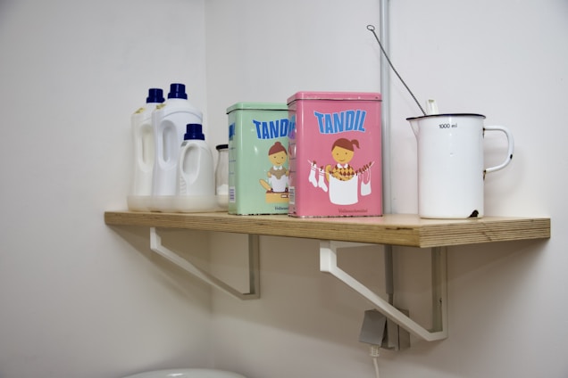 A wooden shelf is mounted on a white wall, holding several items. There are three white plastic bottles with blue caps, likely containing household cleaning products. Beside them, two colorful tins with cartoon images are placed, labeled 'Tandil'. A white enamel jug with a measurement marking of 1000 ml is also on the shelf, accompanied by a metal ladle.
