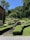 A well-maintained garden with multiple levels of neatly trimmed hedges and a variety of plants. A stone staircase leads up to a higher level where a stone sign reads 'Le Jardin'. In the background, there are people walking and a house partially visible on the left. The surrounding area is lush with greenery and trees.