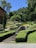 A well-maintained garden with multiple levels of neatly trimmed hedges and a variety of plants. A stone staircase leads up to a higher level where a stone sign reads 'Le Jardin'. In the background, there are people walking and a house partially visible on the left. The surrounding area is lush with greenery and trees.