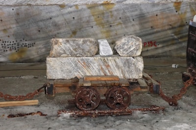 A small, rusted mining cart with two wheels holds several large rough stones. The cart is on a metal track and is connected to a rusted chain. The background is a gray, industrial wall with sporadic markings and patches of discoloration. The ground appears dusty and industrial.