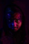 A mysterious portrait featuring a person with dramatic, colorful lighting casting shadows and highlights on their face. The predominant colors are deep blue and pink, creating a moody and striking visual effect. The person appears to be wearing a hood, which adds to the enigmatic atmosphere.