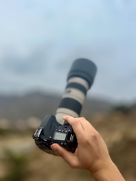 A hand holds a DSLR camera with a long, wide zoom lens. The camera is pointed toward a blurred background suggesting an outdoor setting.