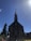 A large stone church with a steep, pointed roof is silhouetted against a clear blue sky. The structure features a prominent bell tower with a cross at the top. People are gathered at the entrance, and tall evergreen trees flank the building.