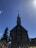 A large stone church with a steep, pointed roof is silhouetted against a clear blue sky. The structure features a prominent bell tower with a cross at the top. People are gathered at the entrance, and tall evergreen trees flank the building.