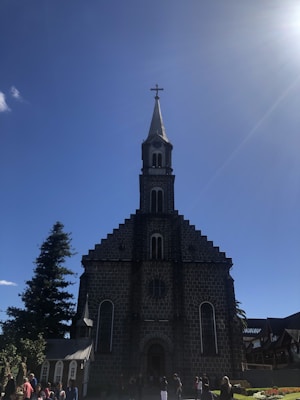 A large stone church with a steep, pointed roof is silhouetted against a clear blue sky. The structure features a prominent bell tower with a cross at the top. People are gathered at the entrance, and tall evergreen trees flank the building.