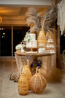 A decorative table is adorned with an elegant, tiered white cake garnished with fresh greenery and flowers. Surrounding the cake are small cupcakes with red and white toppings. Tall, woven lanterns and pampas grass arrangements enhance the ambiance with rustic charm. The setup includes delicate lace accents, complementing the warm, cozy atmosphere.