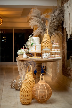 A decorative table is adorned with an elegant, tiered white cake garnished with fresh greenery and flowers. Surrounding the cake are small cupcakes with red and white toppings. Tall, woven lanterns and pampas grass arrangements enhance the ambiance with rustic charm. The setup includes delicate lace accents, complementing the warm, cozy atmosphere.
