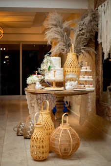 A decorative table is adorned with an elegant, tiered white cake garnished with fresh greenery and flowers. Surrounding the cake are small cupcakes with red and white toppings. Tall, woven lanterns and pampas grass arrangements enhance the ambiance with rustic charm. The setup includes delicate lace accents, complementing the warm, cozy atmosphere.