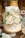 A beautifully decorated white cake stands adorned with elegant white roses and delicate baby's breath flowers. The cake has a rustic frosting style with hints of gold detailing. It sits on a white pedestal, creating a sophisticated and elegant display, with additional floral arrangements and dessert items visible in the background.