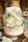 A beautifully decorated white cake stands adorned with elegant white roses and delicate baby's breath flowers. The cake has a rustic frosting style with hints of gold detailing. It sits on a white pedestal, creating a sophisticated and elegant display, with additional floral arrangements and dessert items visible in the background.