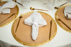 A well-set dining table features woven rattan placemats with a golden decorative edge. White napkins are elegantly folded and placed in the center of each placemat. Gold cutlery is meticulously arranged, with a fork and knife on each side of the placemats. Crystal glasses are set near the top, enhancing the luxurious feel. The tablecloth is light cream, complementing the overall classy setup.