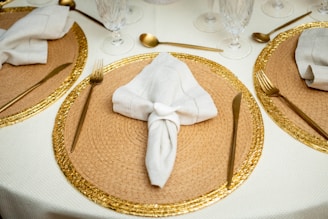 A well-set dining table features woven rattan placemats with a golden decorative edge. White napkins are elegantly folded and placed in the center of each placemat. Gold cutlery is meticulously arranged, with a fork and knife on each side of the placemats. Crystal glasses are set near the top, enhancing the luxurious feel. The tablecloth is light cream, complementing the overall classy setup.