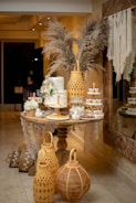 An elegantly decorated dessert table with a multi-tiered white cake adorned with flowers, surrounded by assorted small desserts like cupcakes and pastries. There are decorative elements, including pampas grass in the background and large woven lanterns on the floor. The setting appears to be indoors with a cozy and intimate atmosphere.