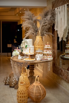 An elegantly decorated dessert table with a multi-tiered white cake adorned with flowers, surrounded by assorted small desserts like cupcakes and pastries. There are decorative elements, including pampas grass in the background and large woven lanterns on the floor. The setting appears to be indoors with a cozy and intimate atmosphere.