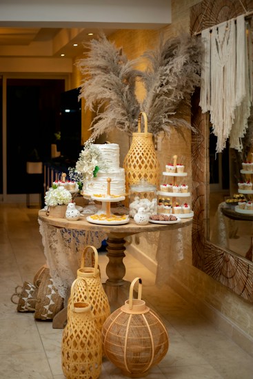 An elegantly decorated dessert table with a multi-tiered white cake adorned with flowers, surrounded by assorted small desserts like cupcakes and pastries. There are decorative elements, including pampas grass in the background and large woven lanterns on the floor. The setting appears to be indoors with a cozy and intimate atmosphere.