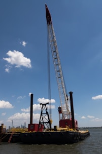 A large barge with heavy machinery, including a tall crane, is floating on a body of water. The crane is painted in red and white, towering above the vessel with a clear blue sky and a few scattered clouds in the background. There are visible red and black structures on the barge, and the water reflects a calm setting.