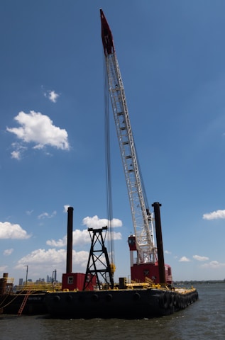 A large barge with heavy machinery, including a tall crane, is floating on a body of water. The crane is painted in red and white, towering above the vessel with a clear blue sky and a few scattered clouds in the background. There are visible red and black structures on the barge, and the water reflects a calm setting.